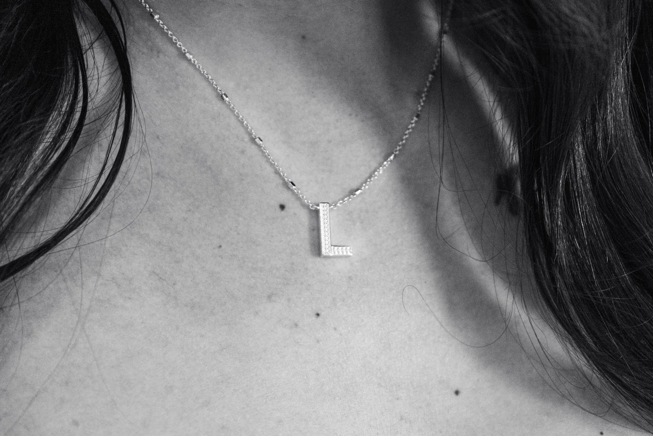 Close-up of a person's neck and chest showing a silver necklace with a pendant in the shape of the letter 'L'.
