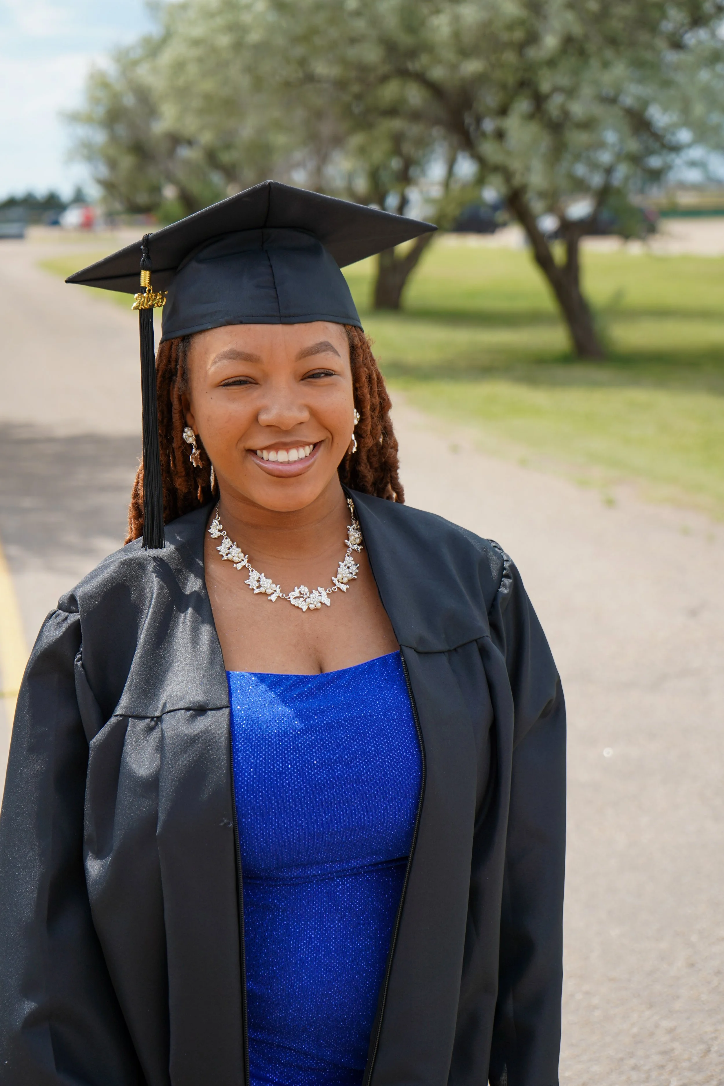 A smiling young woman in a graduation cap and gown stands outdoors on a pathway with trees and grass in the background.