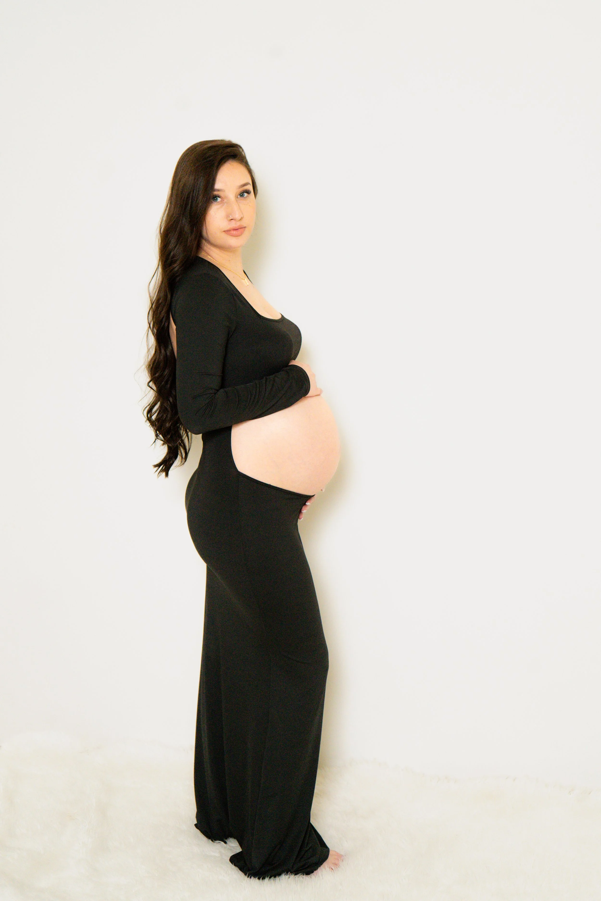 A pregnant woman with long dark wavy hair standing against a plain white wall. She is wearing a black long-sleeve top and black pants, holding her belly with one hand, and looking at the camera.