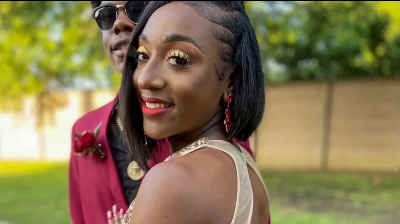 A young woman with dark hair and gold makeup, smiling brightly, standing outdoors next to a man dressed in a red blazer with gold accents, trees, and a wooden fence in the background.