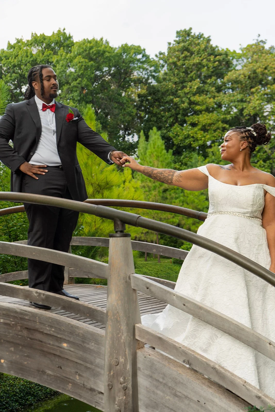 A bride and groom holding hands on a small wooden bridge in a park, with green trees in the background.