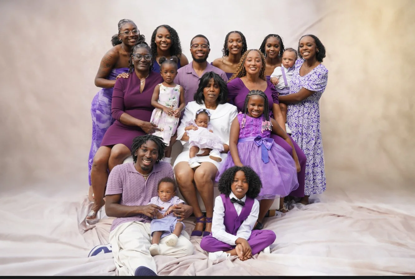 A large family of 15 people, including children and adults, gathered together for a group photo. They are all smiling and dressed in purple and white clothing, with a beige backdrop.