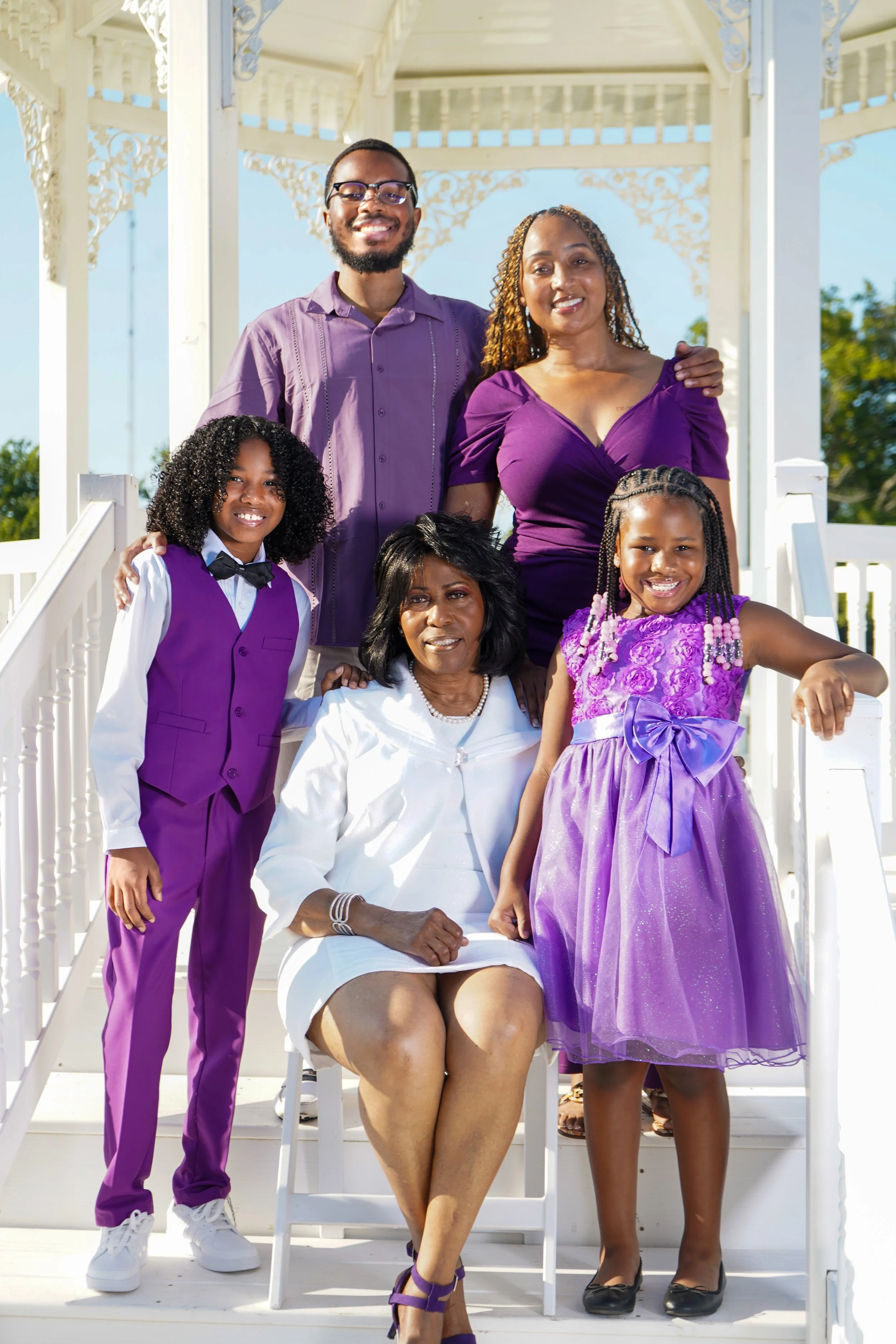 A multi-generational family of six posing on a white gazebo stairs, dressed in purple and white outfits, outdoors on a sunny day.
