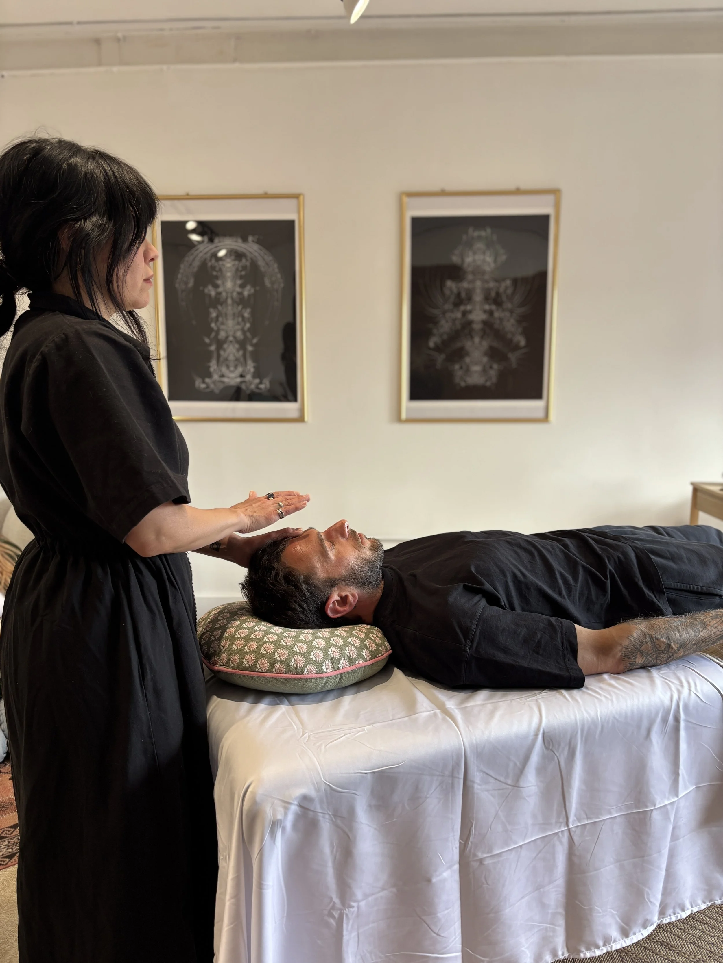 A woman giving a head massage to a man lying on a massage table in a room with framed artwork on the wall.