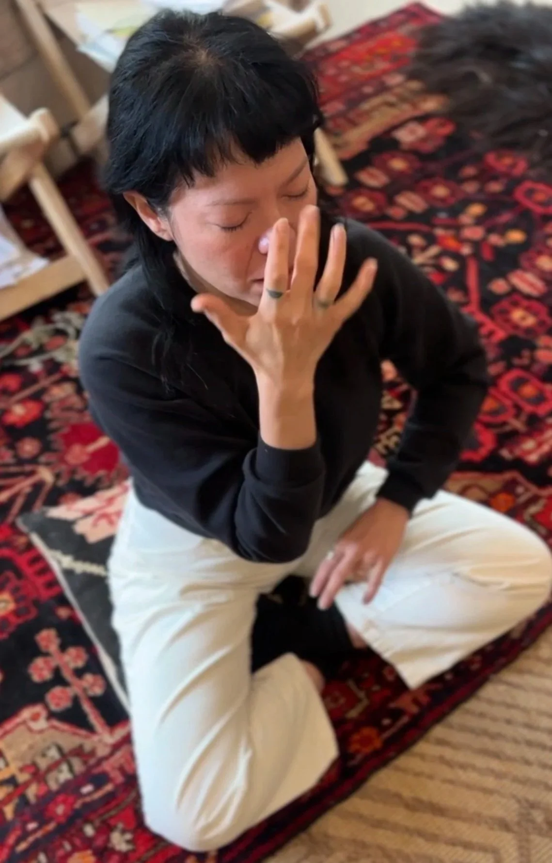 A person sitting on a patterned carpet with their knees crossed, eyes closed, and one hand touching their face in a contemplative pose.