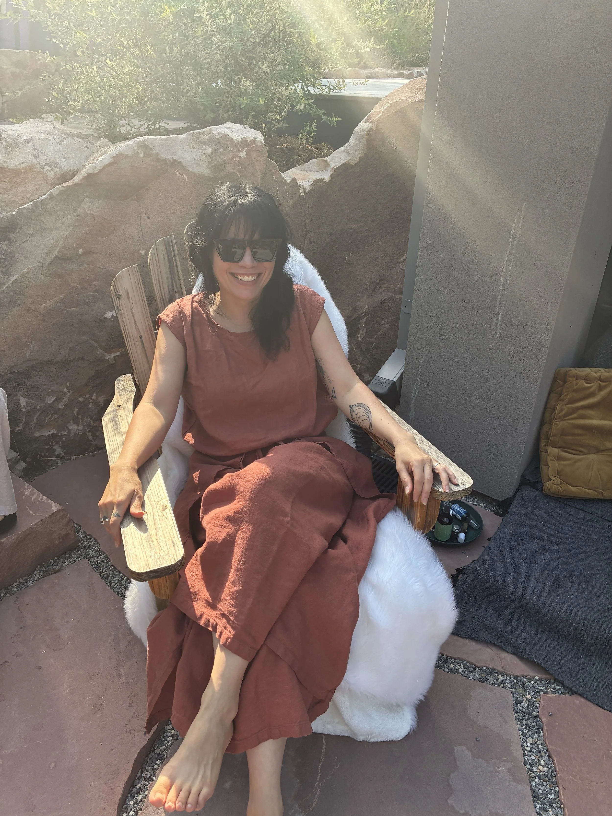 A woman with dark hair wearing sunglasses and a rust-colored dress sitting outdoors on a cushioned wooden chair with a furry white throw, smiling, with rocks and greenery in the background.