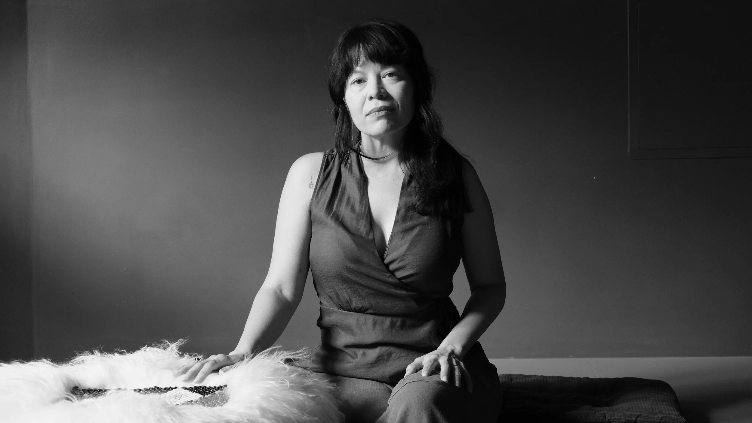 Black and white photo of a woman with dark hair sitting on a cushion, resting her hand on a fluffy blanket, in front of a plain wall.