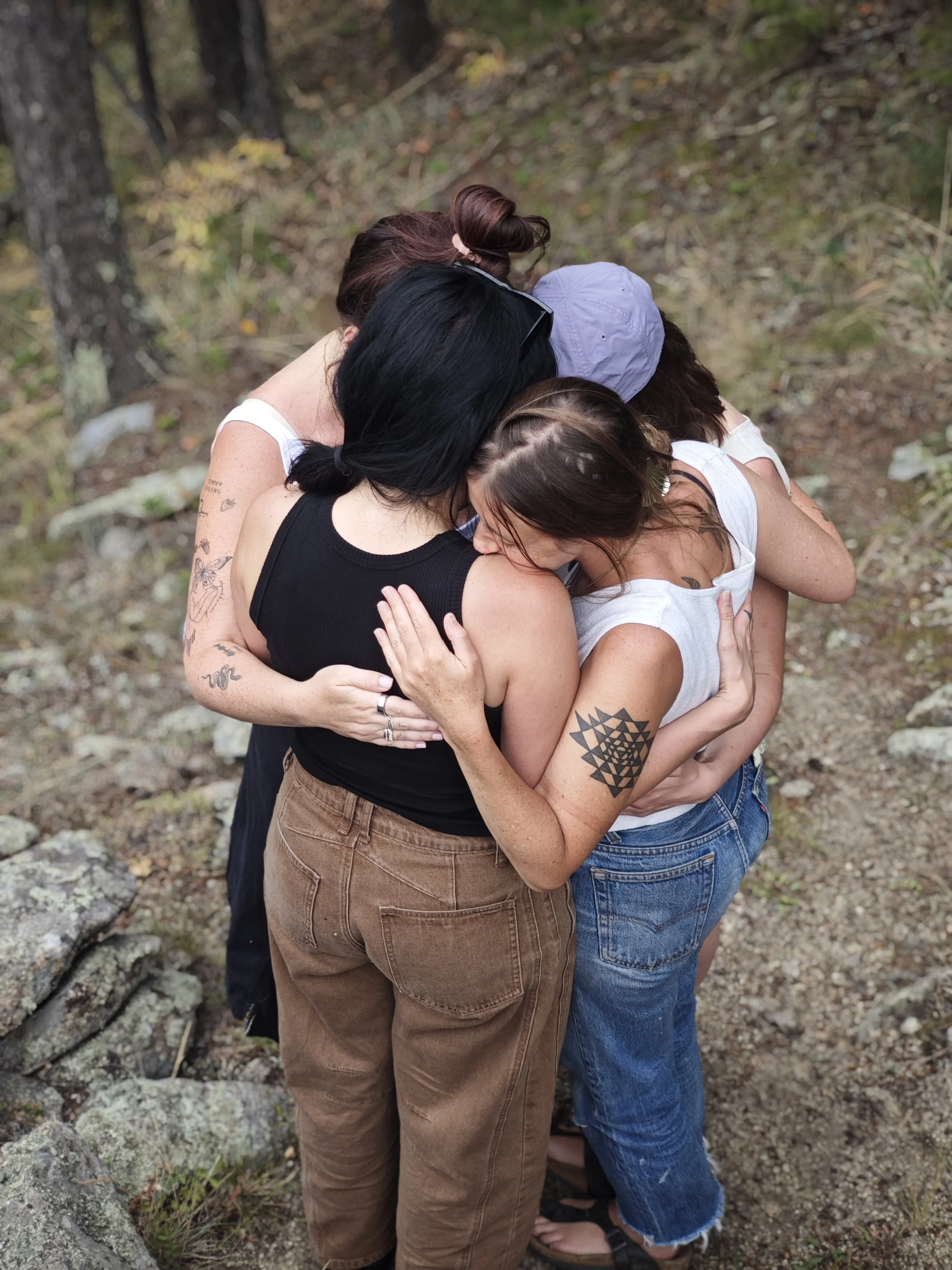 A group of five women embracing in a tight hug outdoors in a wooded area.