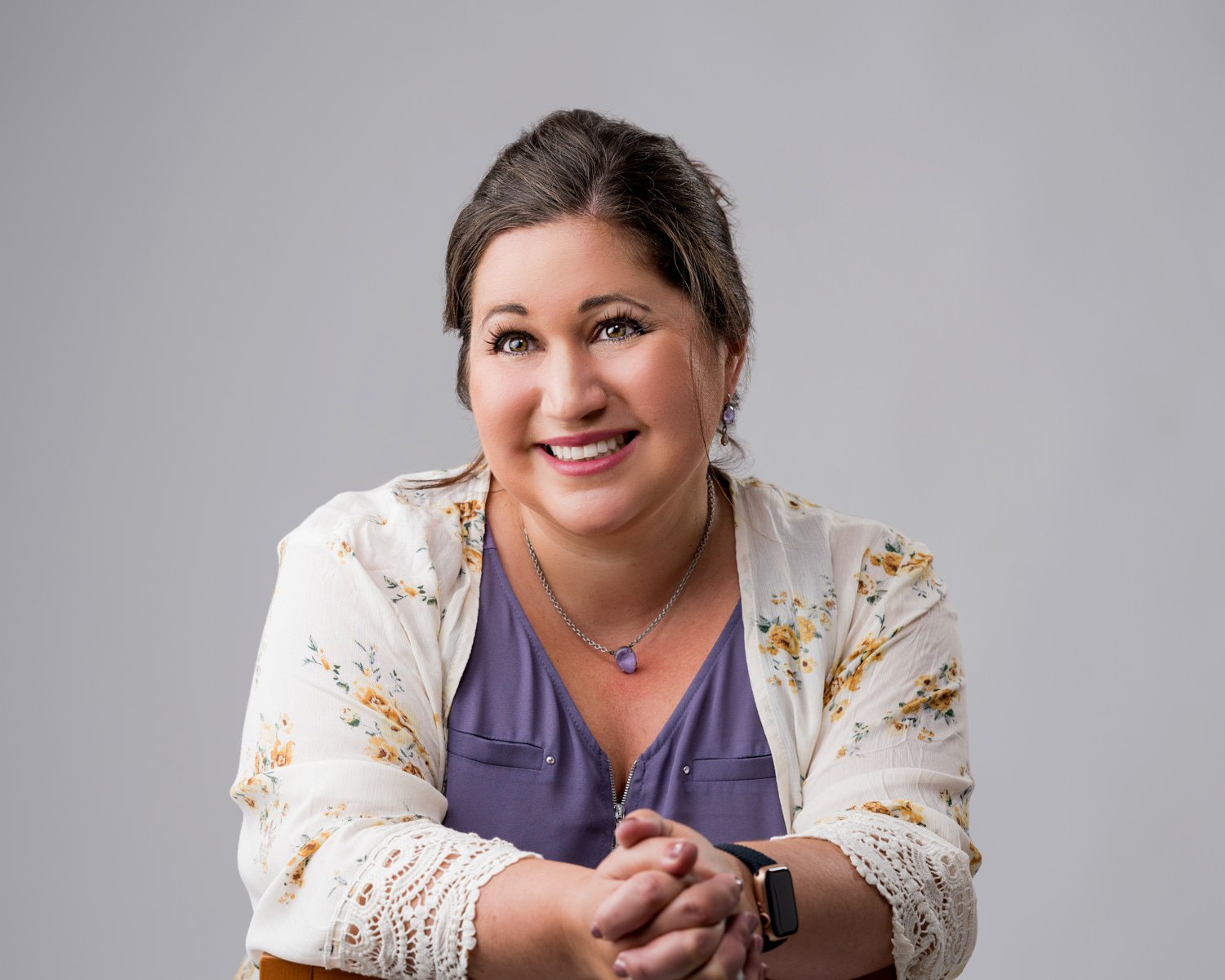 A smiling woman with brown hair and green eyes, wearing a purple top, a white floral cardigan, the necklace and earrings, with her hands clasped, against a gray background.