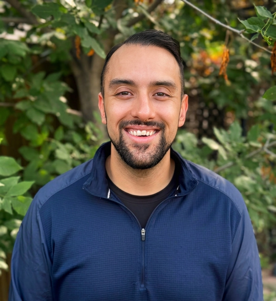 A smiling man with dark hair, a beard, and mustache, wearing a blue zip-up jacket, standing outdoors in front of green foliage.