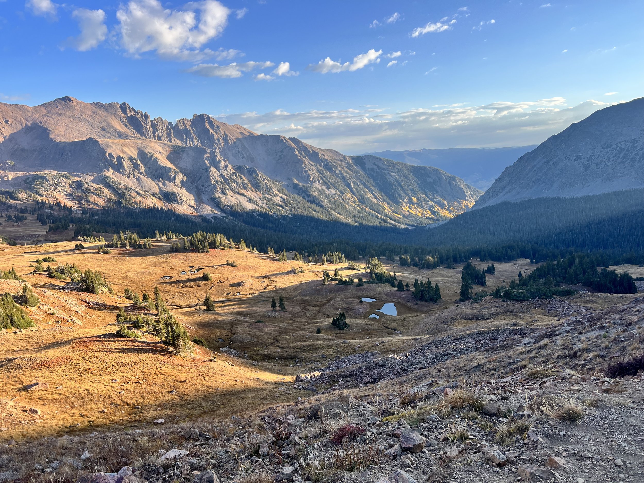 A scenic mountain landscape with rugged peaks, rolling valleys, patches of green forests, small lakes, and a partly cloudy sky.