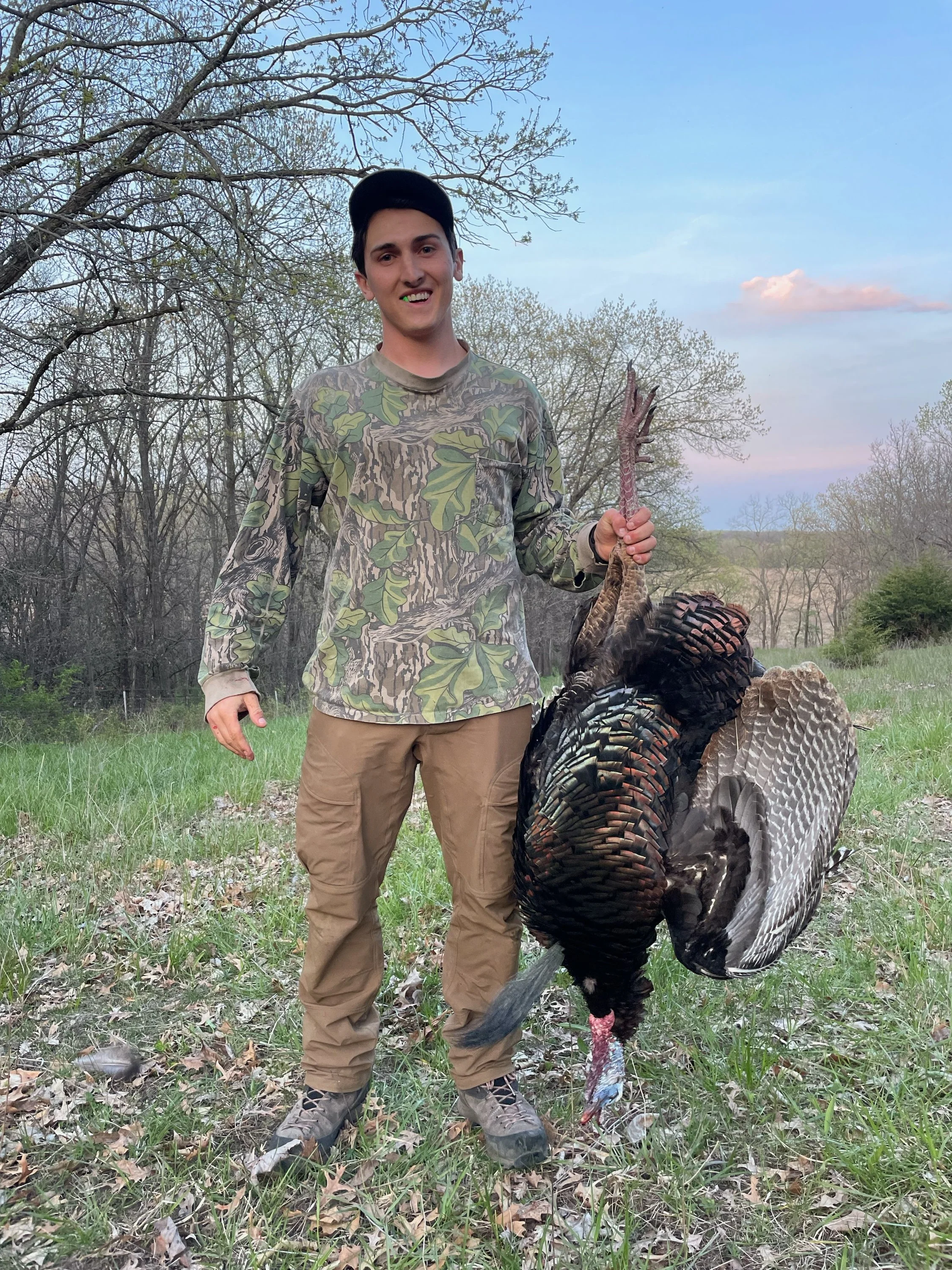 A young man in camouflage clothing holding a large turkey outside in a forested area during evening time.