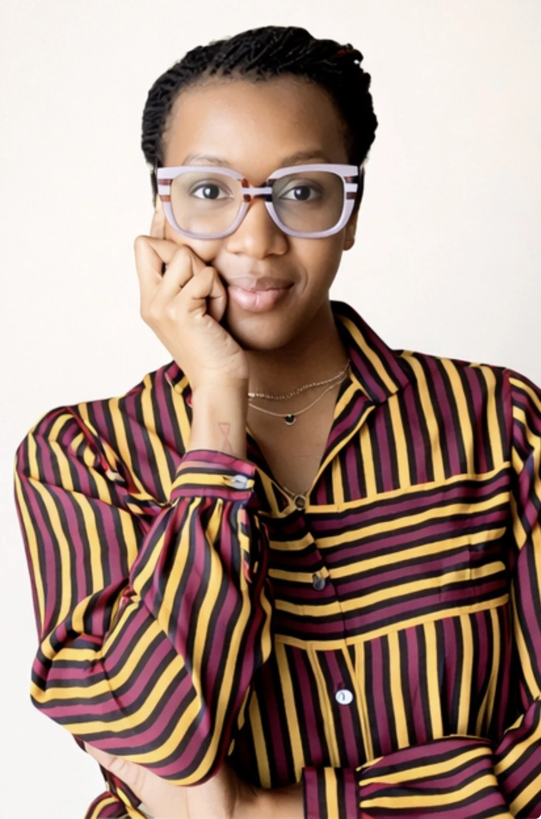 A woman with braided short hair wearing purple glasses and a colorful striped shirt, standing against a white background.