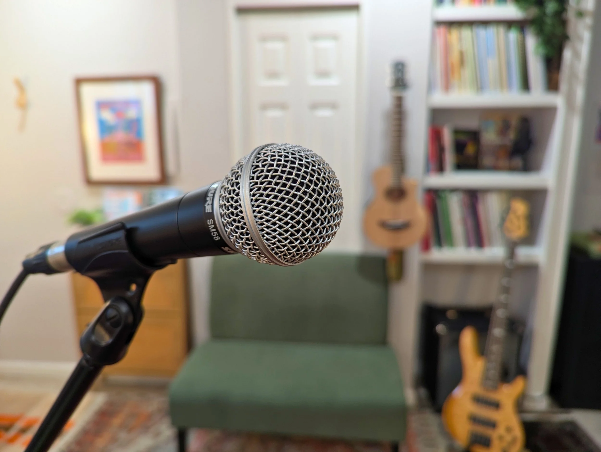 A microphone on a stand in the foreground with a voice studio setup in the background, including a guitar and bookshelf.