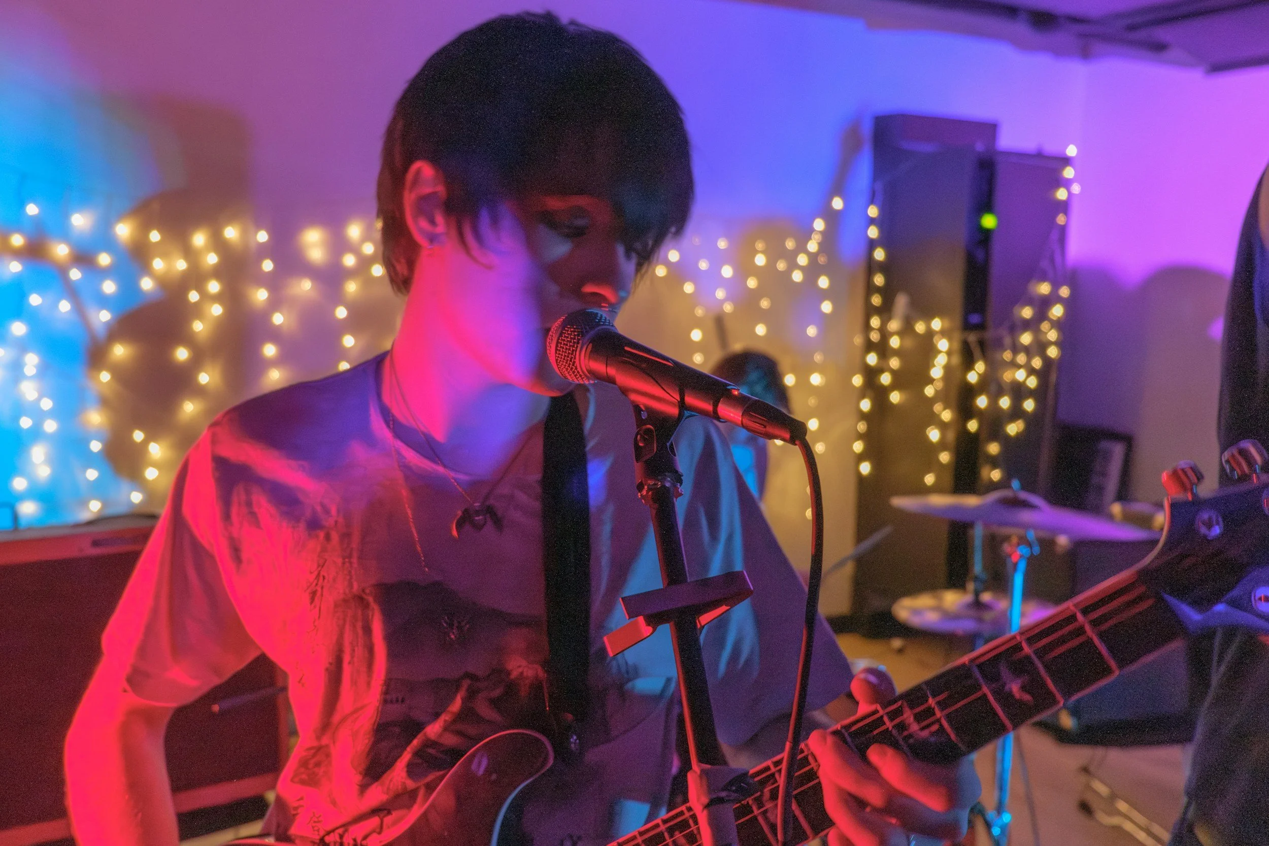 A young nonbinary singer and songwriter with short dark hair singing into a microphone while playing an electric guitar in a dimly lit room decorated with colored lights and string lights in the background.