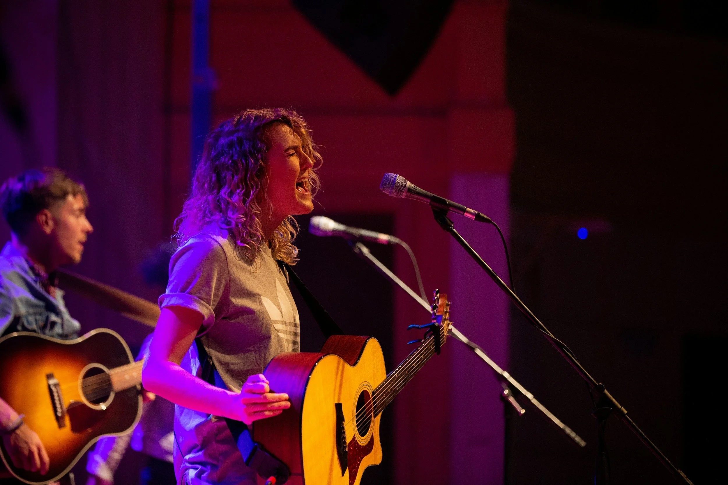 A woman with curly hair singing into a microphone while playing an acoustic guitar on stage, with a man in the background also playing guitar.