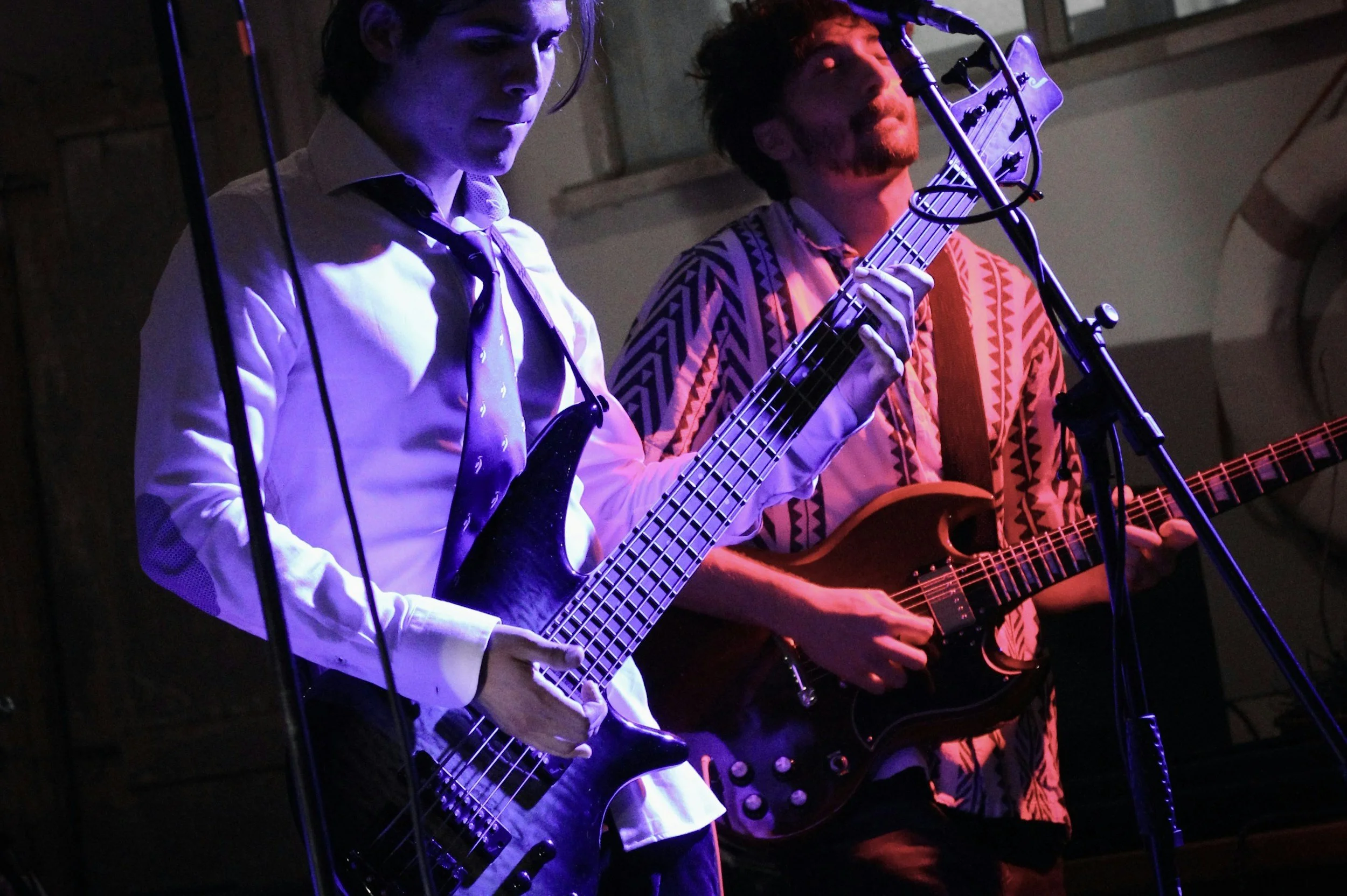 Two musicians playing electric guitars on stage, one wearing a white shirt and tie, the other wearing a patterned shirt, under purple stage lighting.