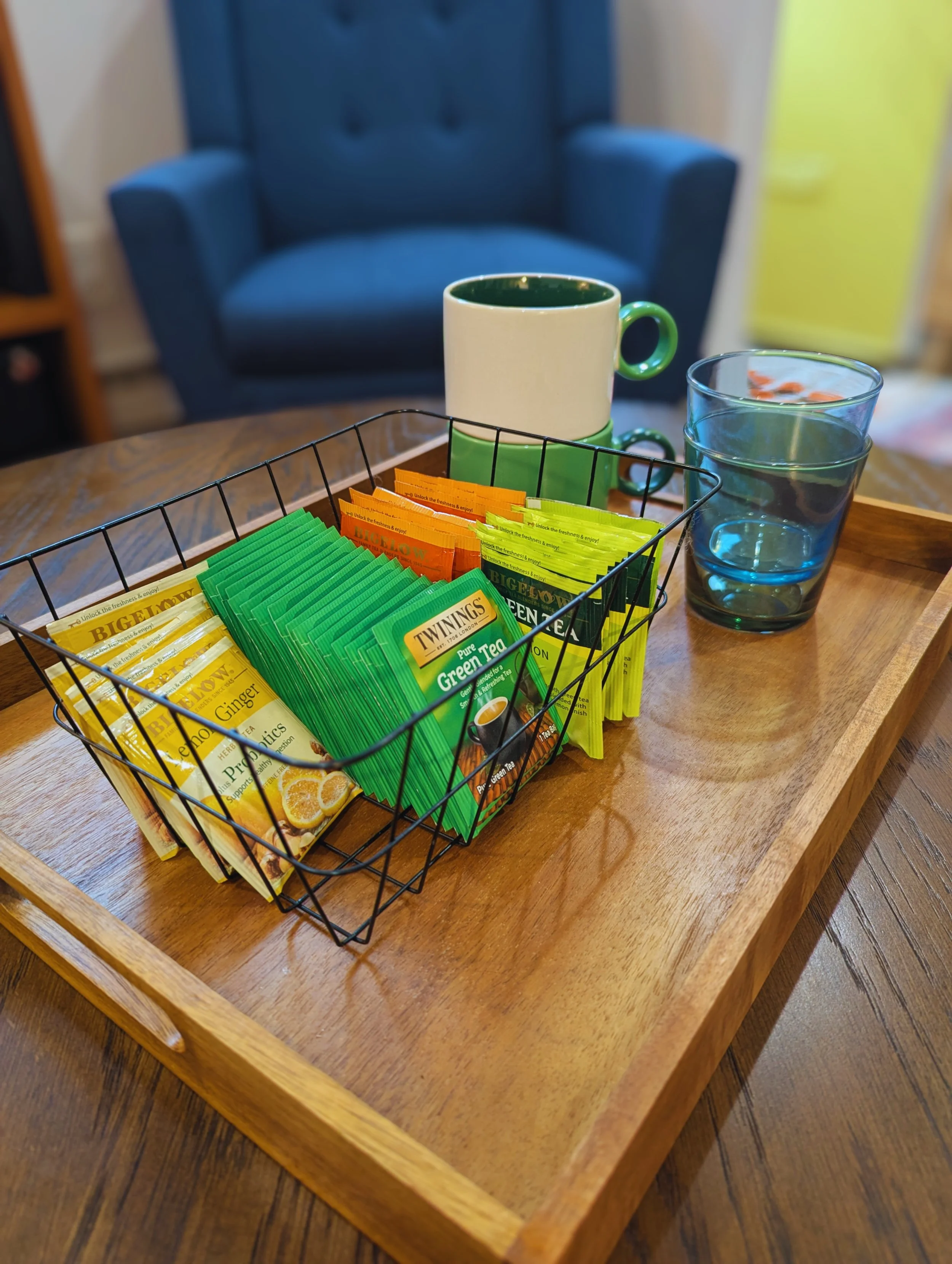 A wooden tray with a wire basket containing assorted tea bags, a white mug with green interior, two glasses of water, and a blue armchair in the background.