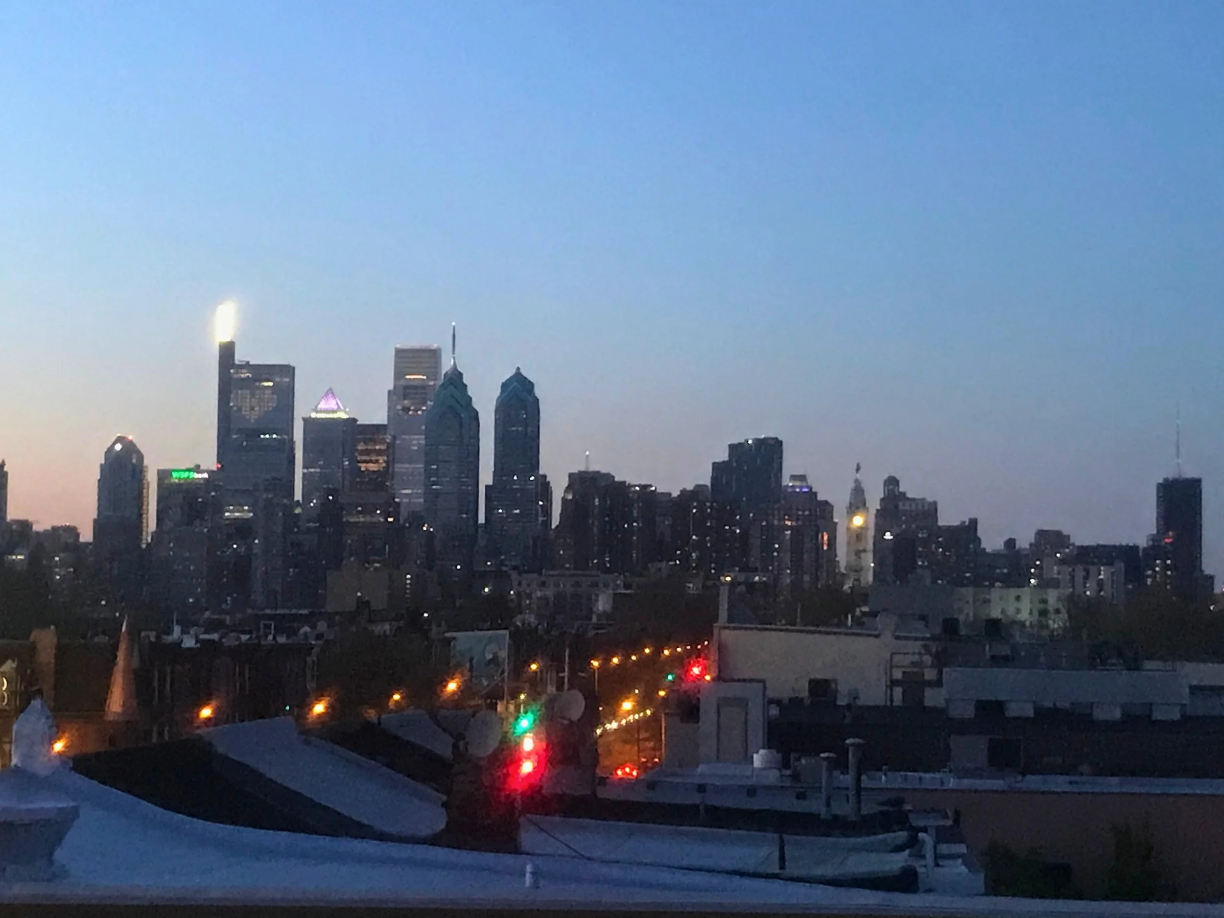 View of the Philadelphia city skyline at dusk with tall skyscrapers and a few lit buildings, with rooftops and city streets in the foreground.