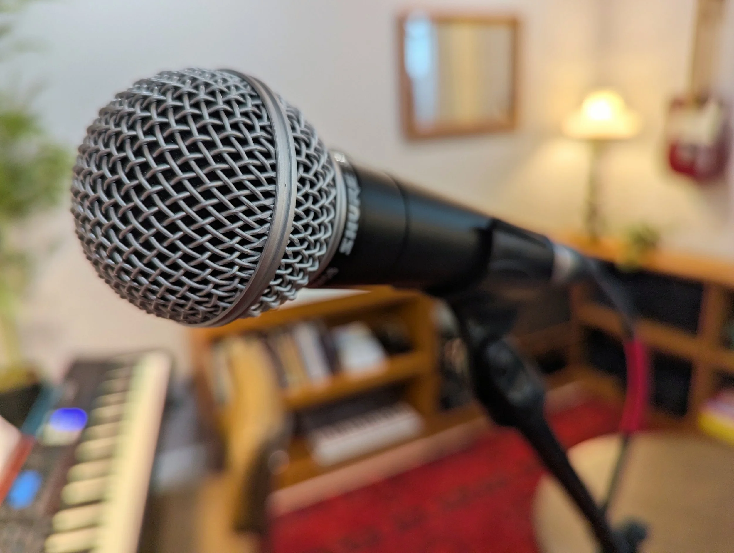 Close-up of a handheld microphone in a home music studio with a keyboard, bookshelf, and lamp in the background.