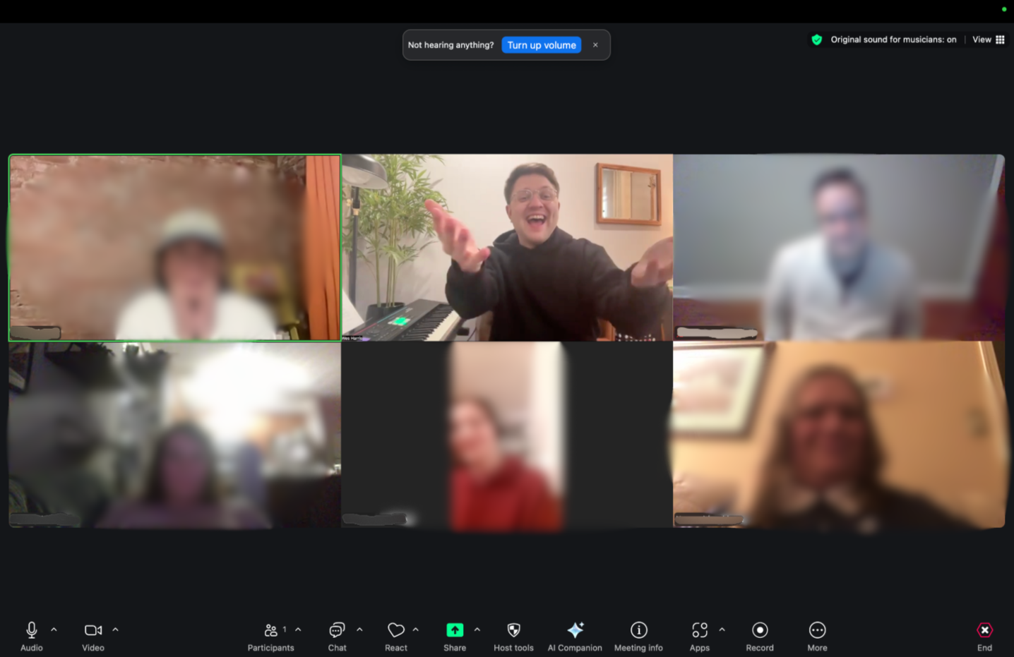 Zoom video call with six participants, Wes Harris, studio owner and voice coach, is smiling and gesturing, with a keyboard and plants in the background, others are blurred an engaged in the voice group class.
