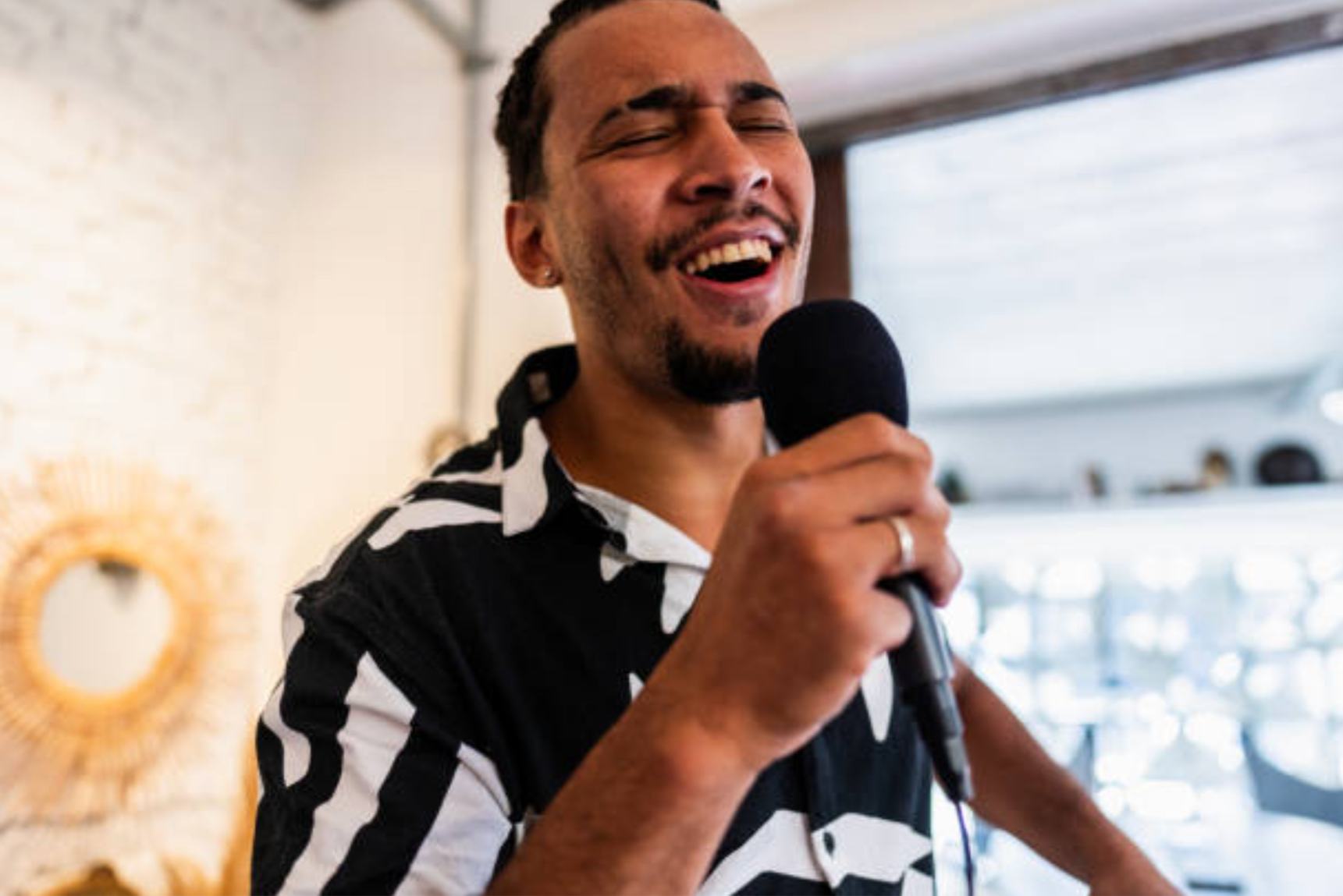 A man singing or speaking into a microphone, smiling with eyes closed, in an indoor setting with a large window behind him.