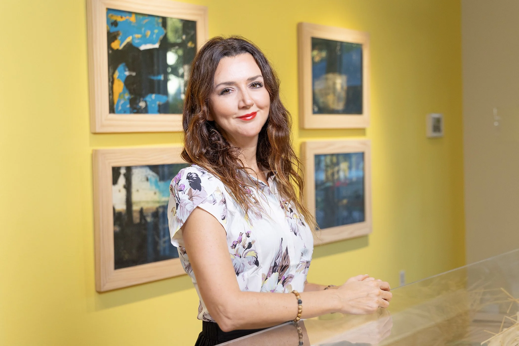 A woman with wavy brown hair, wearing a floral blouse, stands in an art gallery with yellow walls and four framed abstract paintings in the background.