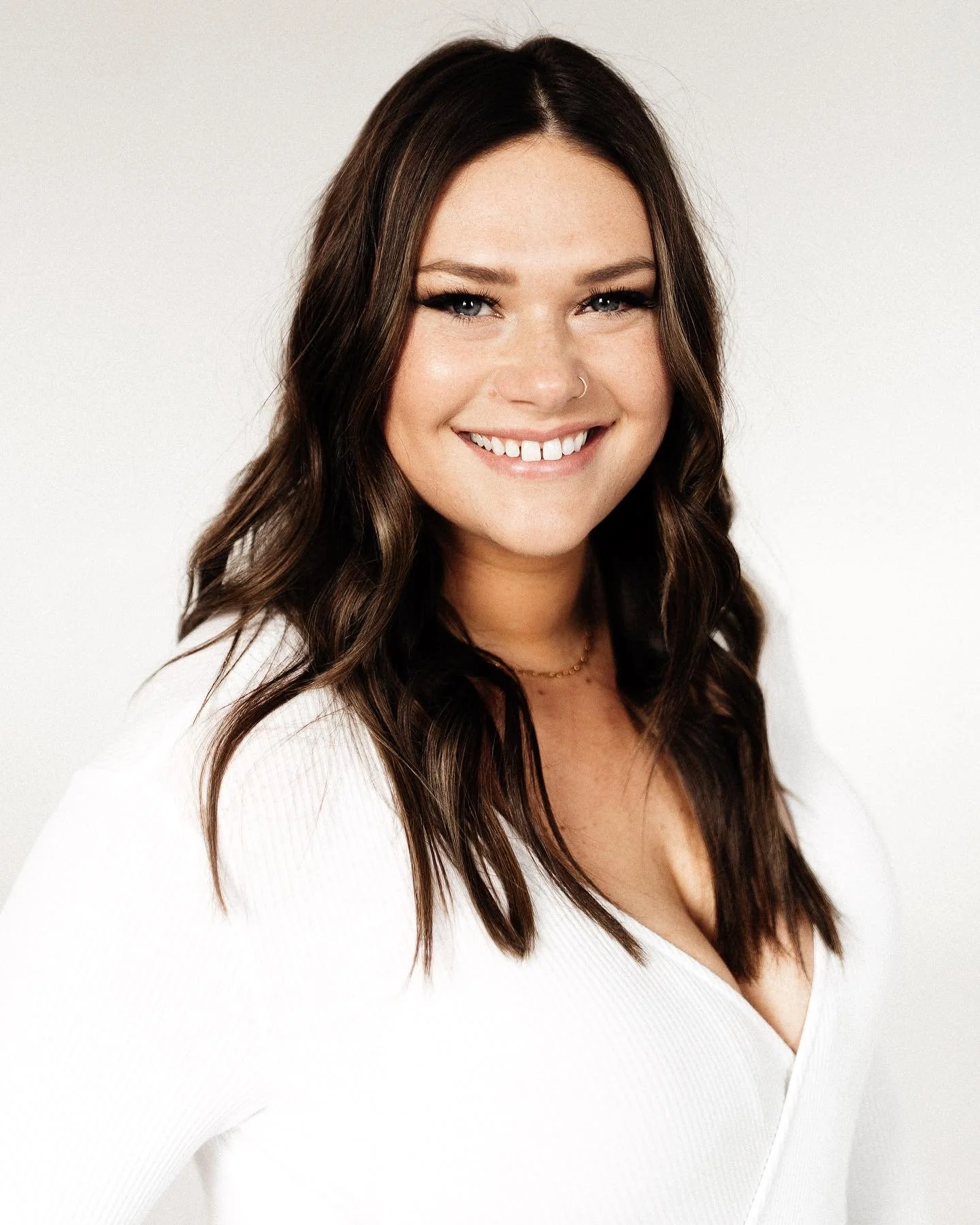 A woman with long, wavy brown hair smiling at the camera, wearing a white top and a small gold necklace, against a plain white background.