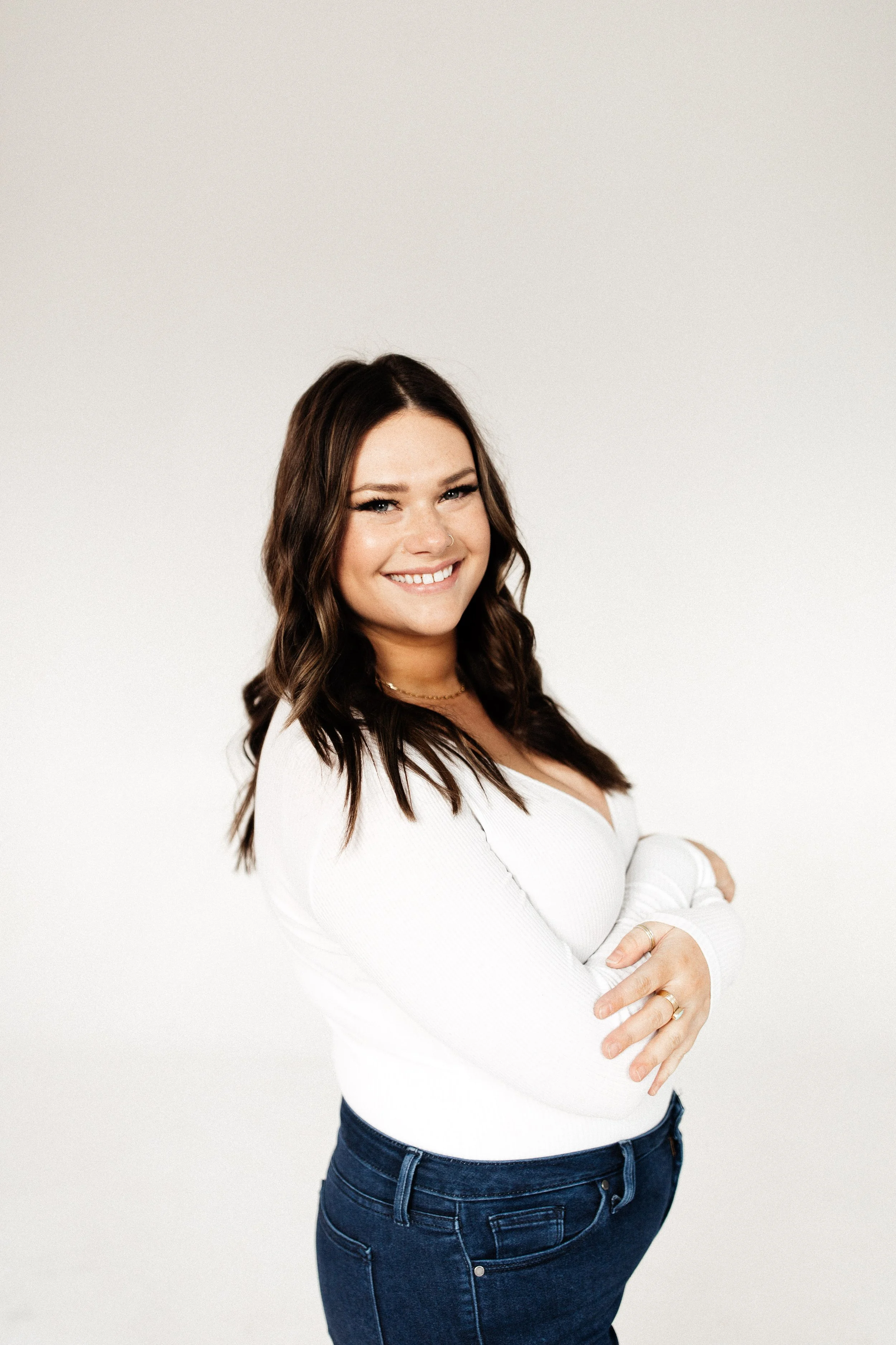 A woman with dark brown hair, smiling, wearing a white long-sleeve sweater and dark blue jeans, standing against a plain white background.