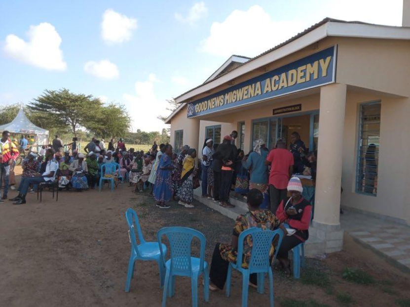 Crowd of people outside and in front of a building labeled 'Good News Migwena Academy', with some sitting on blue plastic chairs and others lining up at windows inside the building.