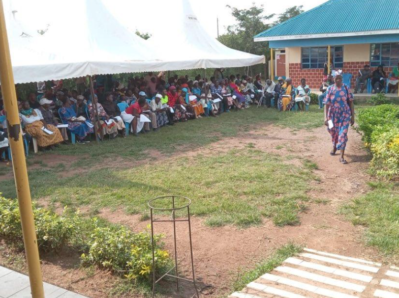 A large gathering of people, mostly women, seated under a white tent and in chairs outside a building, with a woman walking along a dirt path nearby in a rural setting.