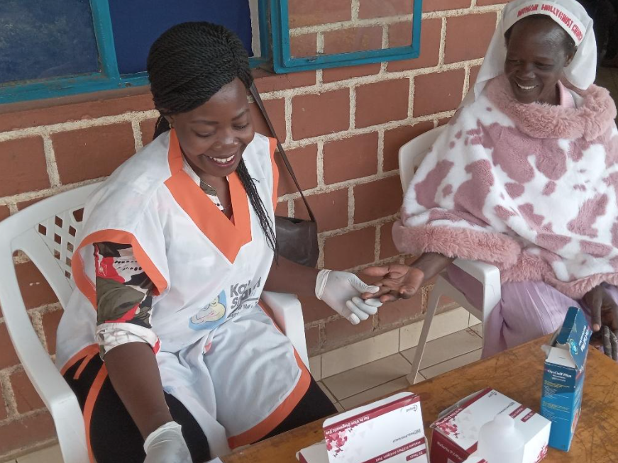 A healthcare worker in a white and orange uniform is seated, smiling, and holding an elderly woman's hand. The elderly woman is wearing a pink and white patterned blanket and a head covering, also smiling. They are outside against a brick wall, and there are medical supplies on the table in front of them.