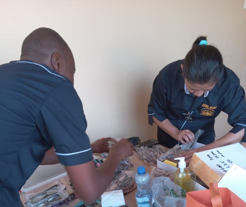 Two people in dark blue uniforms working at a table with small plastic bags, bottles, and a box in a room with plain white walls.