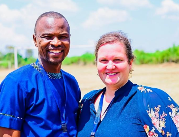 A man and a woman standing outside, smiling for the camera with clear skies and green landscape in the background.