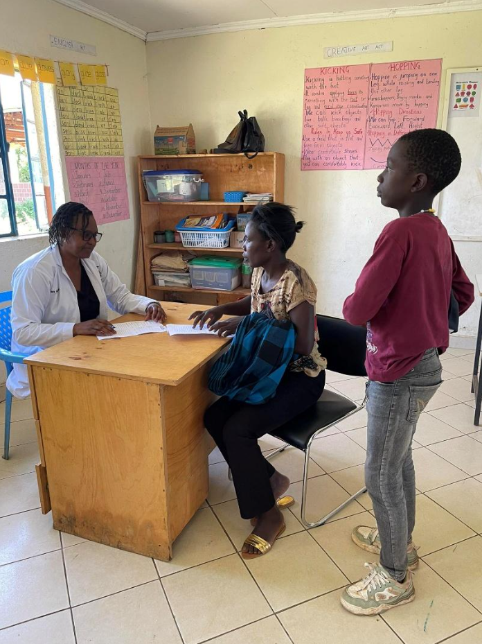 A woman sits at a desk with a medical professional, talking. A female patient sits across from the woman, and a young girl stands nearby observing. The room has posters on the wall, a shelf with supplies, and a window.