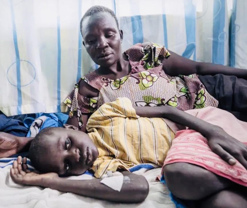 A woman and a young child lying on a bed in a room with striped curtains. The woman is sitting up and looking at the child, who is lying down with their head resting on a pillow and arm bent under their head.