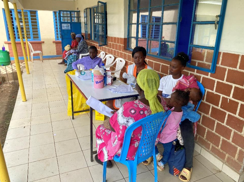 A healthcare setting with women and children seated at a table, engaging with healthcare workers. There are windows with blue frames, a brick wall, and chairs along the wall.
