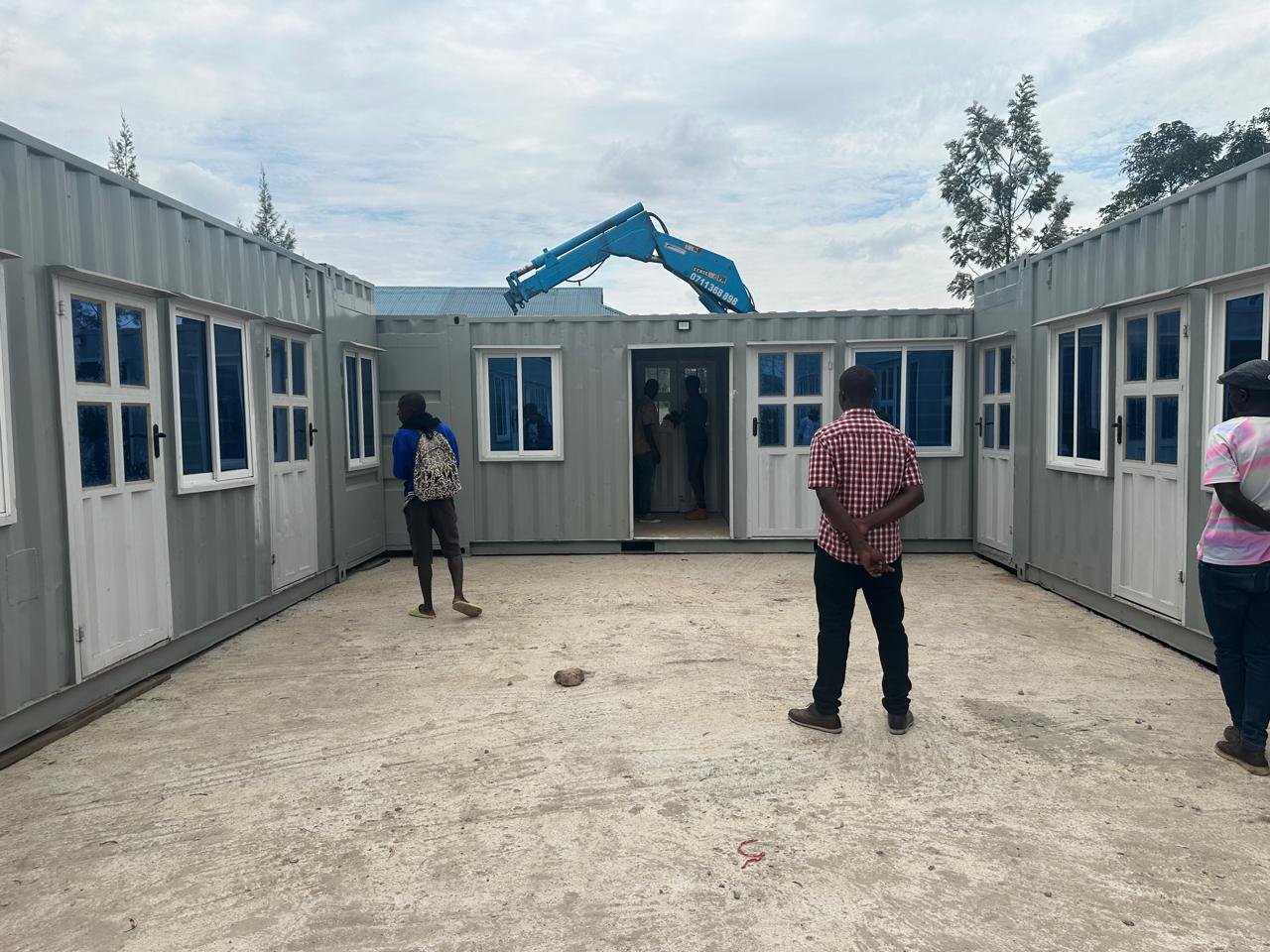 Three people are standing in a courtyard surrounded by three shipping containers.