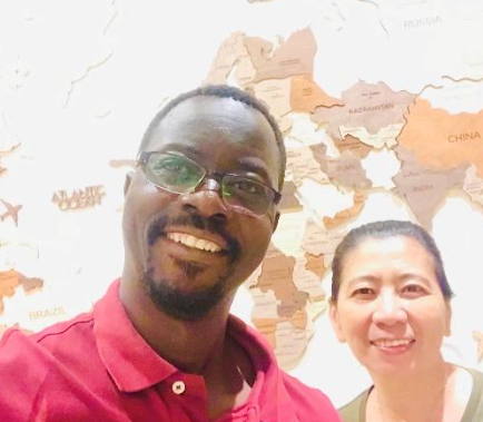 A smiling man with glasses and a beard wearing a red shirt taking a selfie with a smiling woman with dark hair pulled back, standing in front of a world map on the wall.