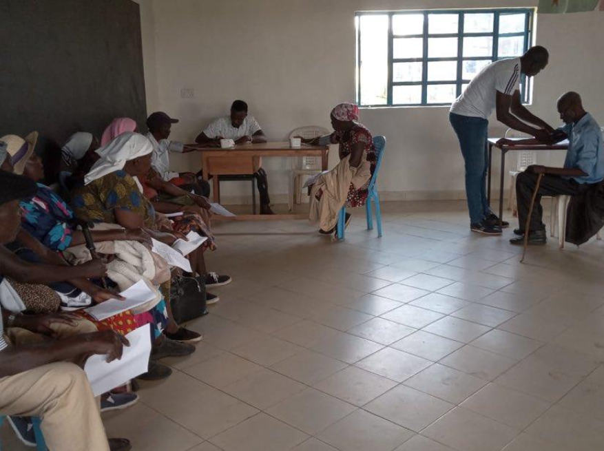 A group of people seated on chairs on the left side of a room, some holding papers, while two men are standing near a seated woman on the right, receiving medical care inside a room with a window on the wall.