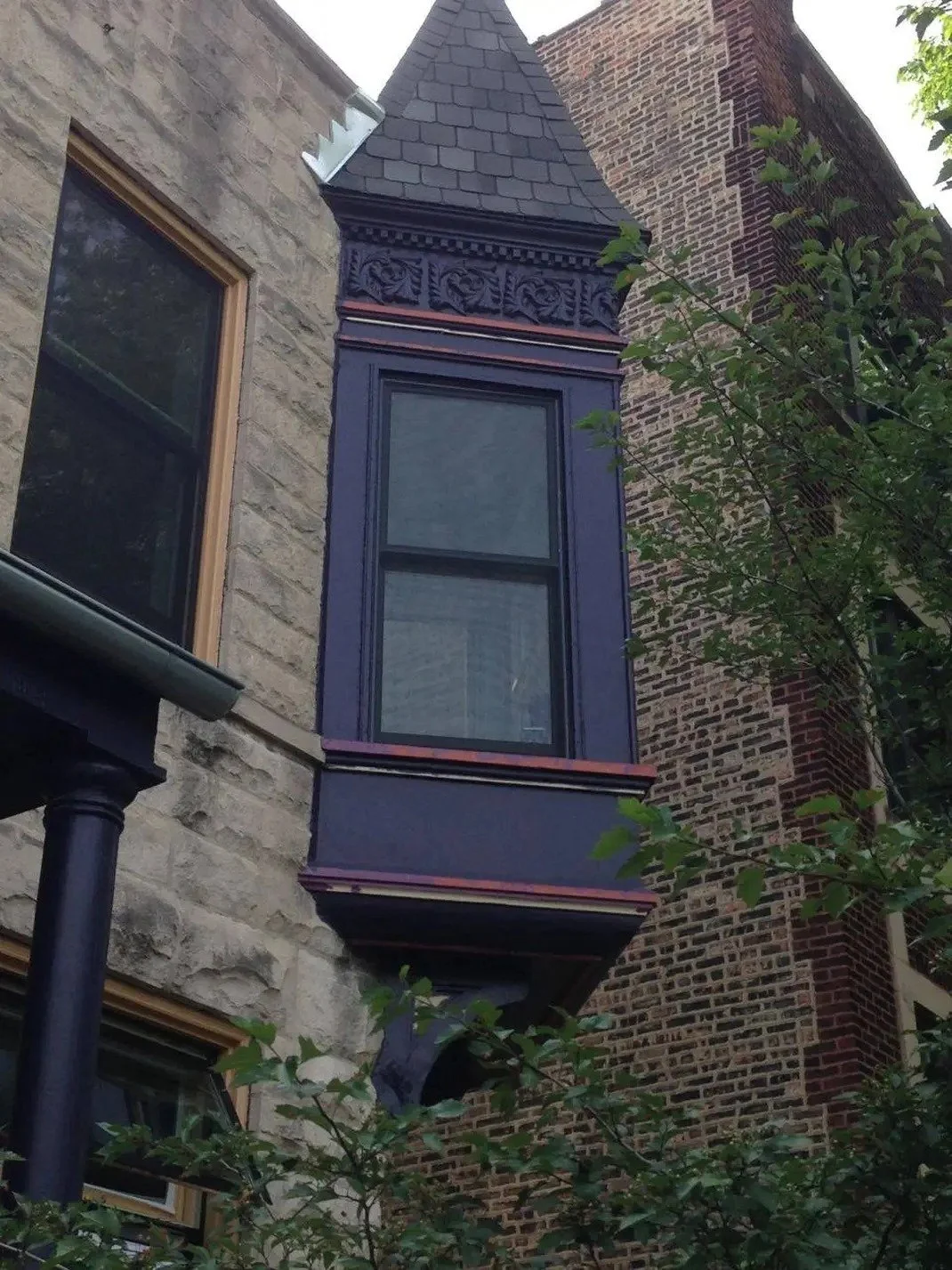 A Victorian-style bay window with ornate trim facade featuring a large window previous paint scheme prior to restoration, a stone exterior, a brick chimney, and greenery front.