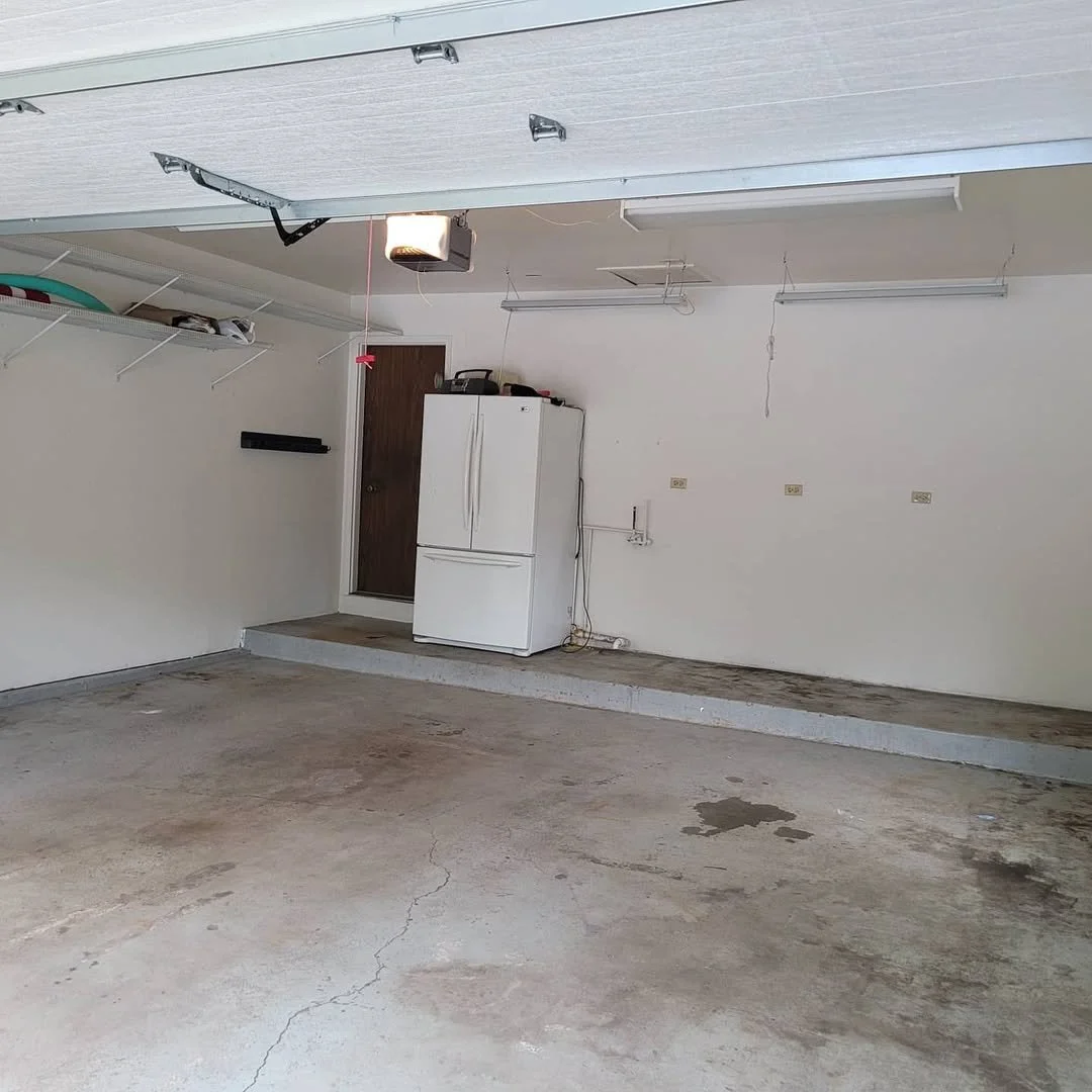 Empty garage with stained concrete floor before sealing with epoxy, white walls, a white refrigerator, and a brown door. Garage ceiling has exposed lighting fixtures and a garage door opener.