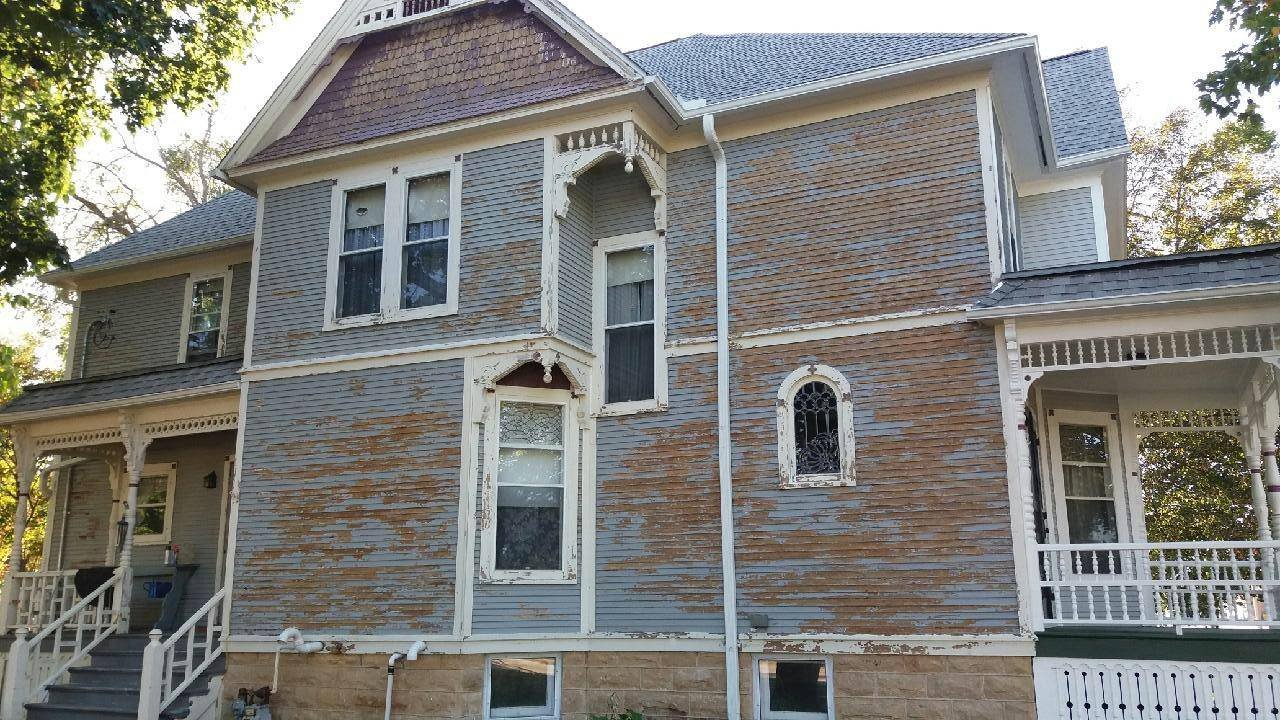 A large Victorian-style house with peeling paint and weathered siding, featuring decorative trim and a porch with white railings.