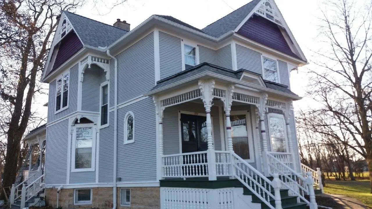 A large Victorian-style house painted in light blue with white trim, featuring decorative woodwork and a wraparound porch, set in a yard with trees.