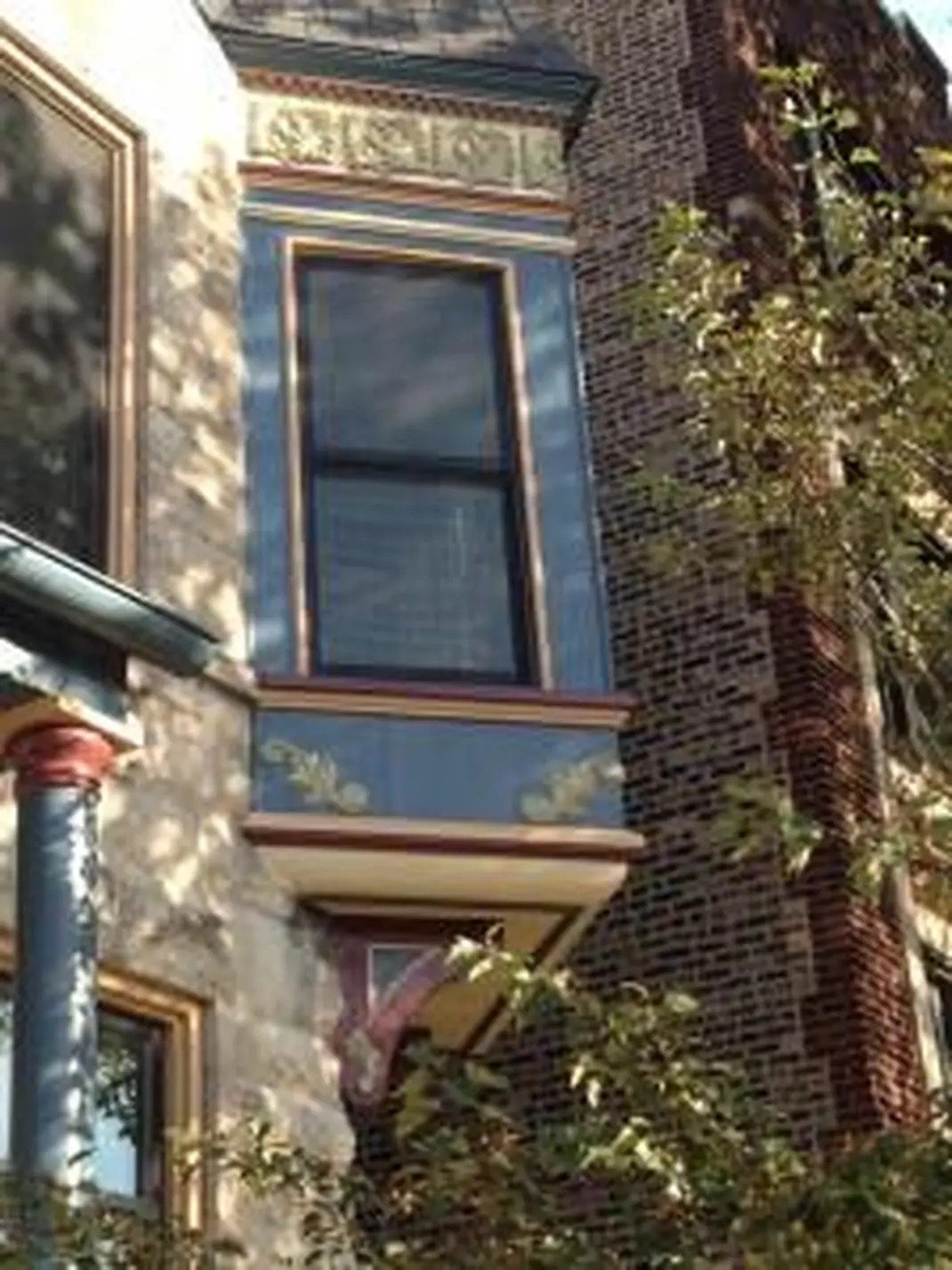 A restored to original paint color Victorian-style bay window with ornate trim building facade featuring a large window with bronze trim, a stone exterior, a brick chimney, and greenery in front.