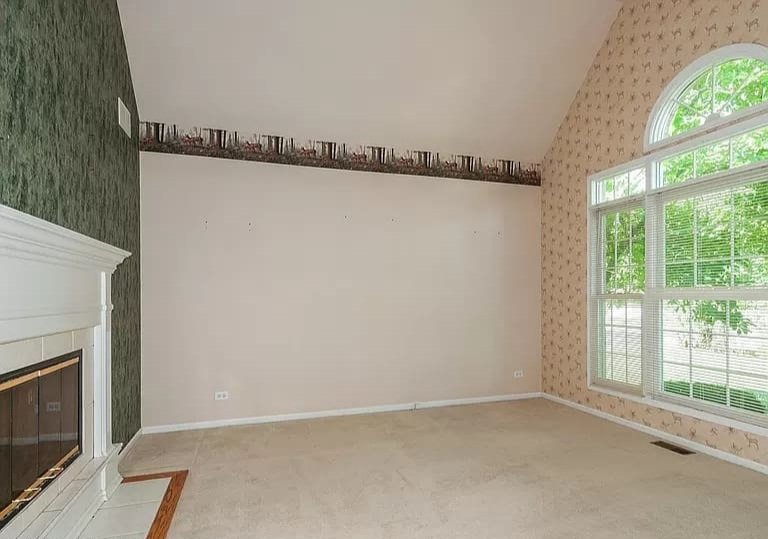 Living room with three different wall paper patterns, fireplace, large arched window with blinds, and beige carpet.