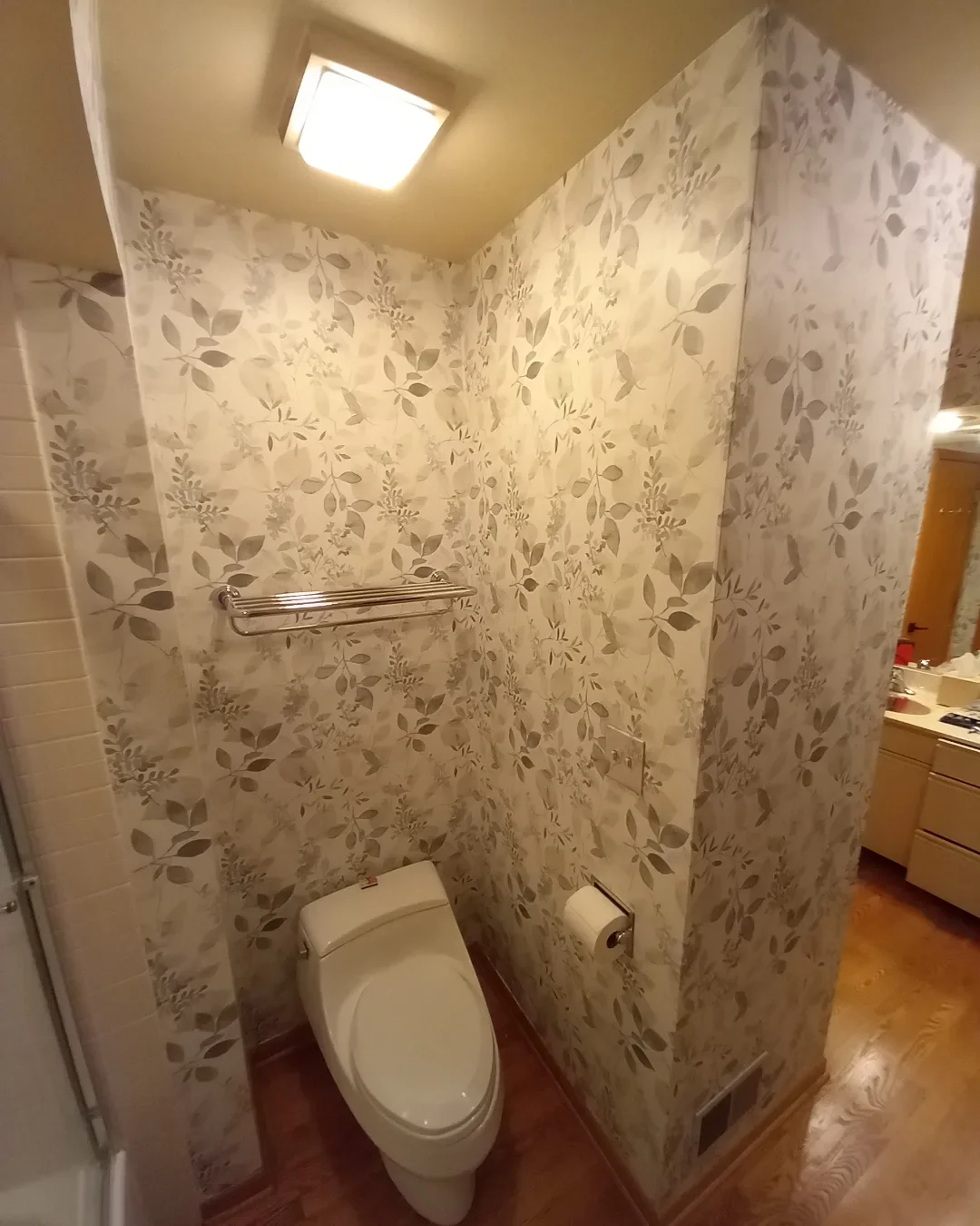Small bathroom with floral wallpaper, toilet, toilet paper, towel rack, ceiling light, and partially visible shower and vanity area.