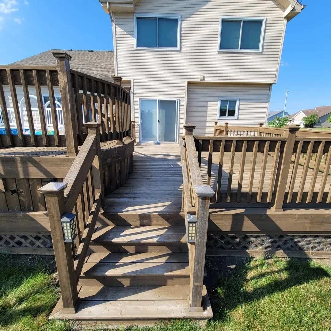 A wooden deck with stairs prior to repainting, leading up to a house in a suburban neighborhood, under a bright blue sky.