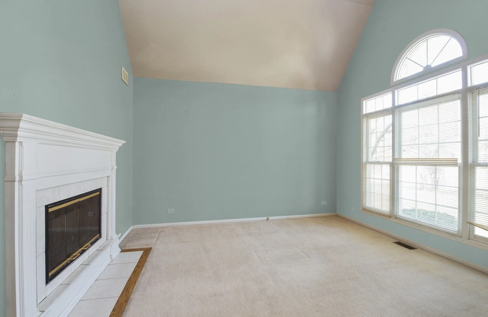 Empty living room with wall paper removed and walls painted light blue,  large bay window, a white fireplace, beige carpet, and high vaulted ceiling.