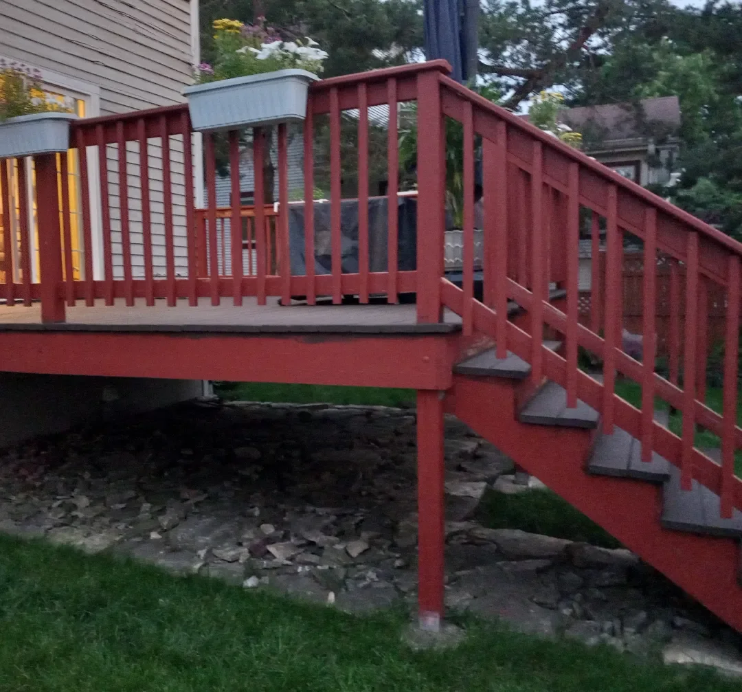 An outdoor wooden deck with red railings and gray steps, attached to a house, with potted plants and flowers, in a backyard with trees and neighboring houses visible in the background.